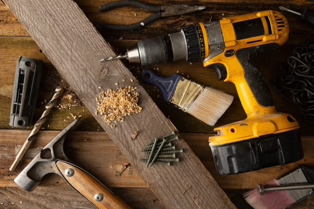 Cordless drill, hammer, paintbrushes, pliers, drill bits, screws and sawdust on a weathered wood workbench