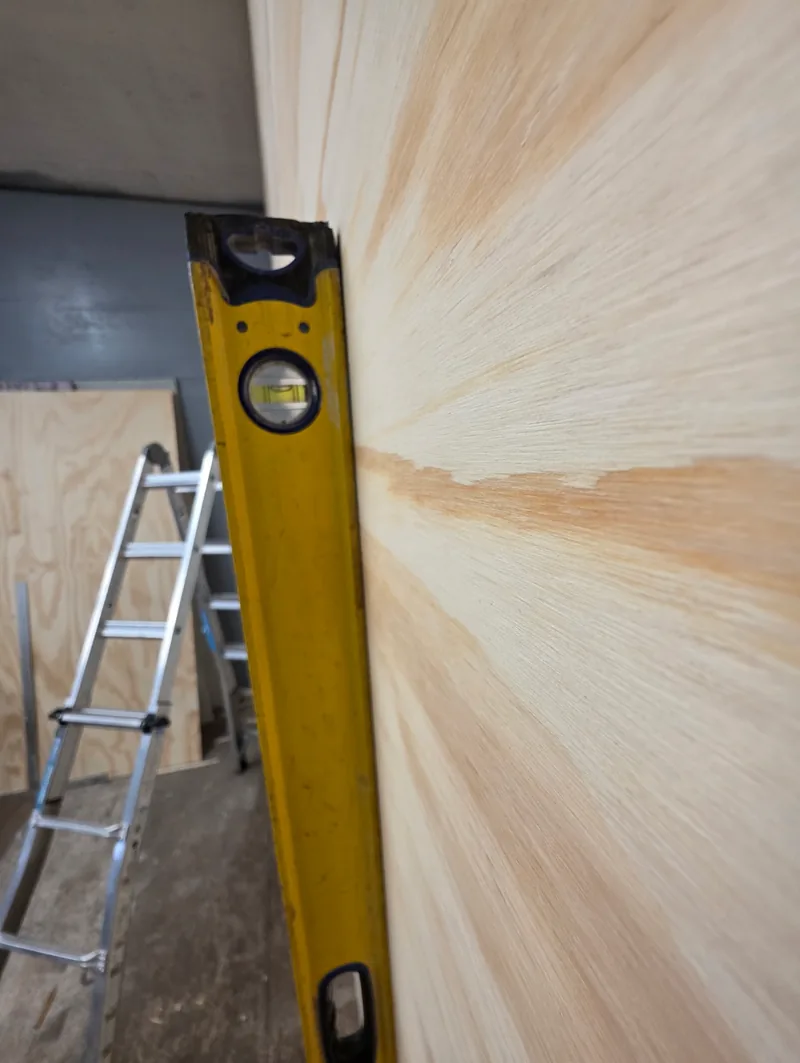 Level held against a new plywood wall to check it is plumb, with a ladder in the background
