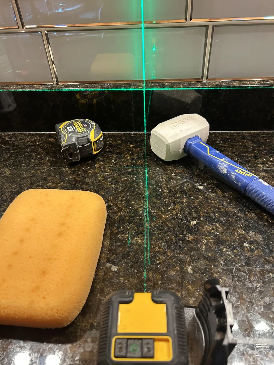 Laser level projecting green lines onto a subway tile backsplash and granite countertop, with a tape measure, rubber mallet and grout sponge